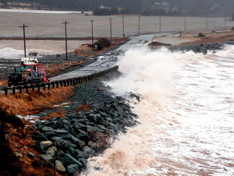 A flooded, partially damaged oceanside road with a wave crashing onto it. 