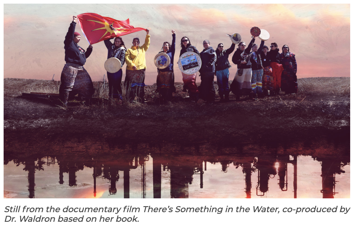 Text reads "Still from the documentary film There's Something in the Water, co-produced by Dr. Waldron based on her book." Above is an image of a group of people standing by a river, many of whom are wearing ribbon skirts. Some hold up intricately decorated drums, and several are holding aloft a First Nations flag. The reflection in the water is of an industrial plant.