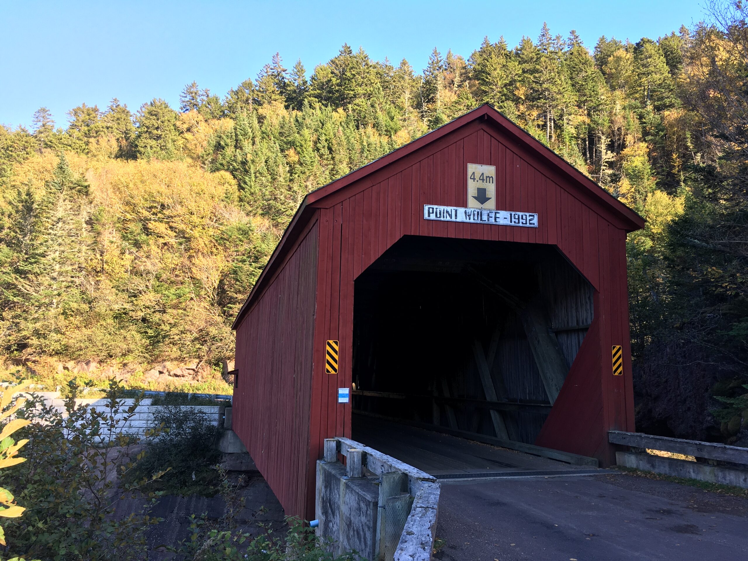  A red covered bridge in front of a hill with trees. The sign on the bridge reads "Point Wolfe-1992". 