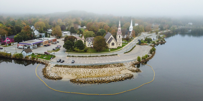 The shoreline of a small town is pictured. In the centre is a wider area of shoreline that has plants growing in the sand, and is bordered by a yellow fence in the water.