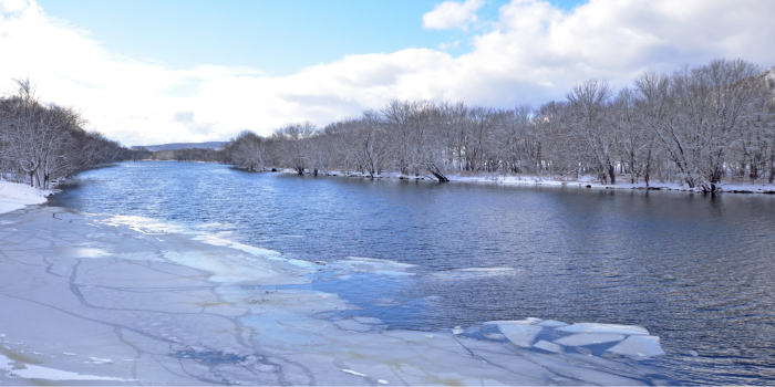 A river scene in winter, with ice covering part of the water and snow on the trees and ground.