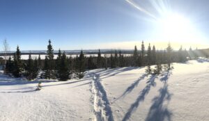 An image taken from the top of a snow covered hill. Trees and a body of water are seen in the distance. There is a trail of footprints through the snow and the sun is shining.