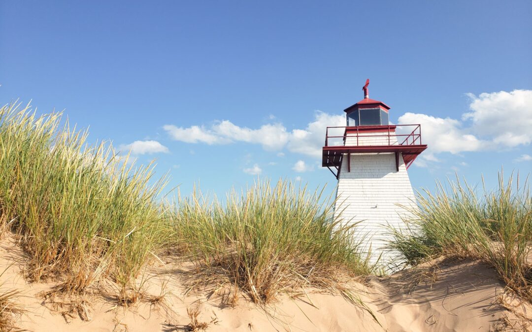 Le français suit. A white lighthouse, with red trim, seen from below a sand dune on which green grasses are growing. The sky is blue with white fluffy clouds. Un phare blanc, bordé de rouge, vu de dessous une dune de sable sur laquelle poussent des herbes vertes. Le ciel est bleu avec des nuages blancs et cotonneux.