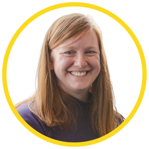Le français suit. A picture of a person with long ginger hair wearing a dark top smiling at the camera, framed by a yellow circle.Photo d'une personne aux longs cheveux roux, vêtue d'un haut sombre, souriant à l'appareil photo, encadrée d'un cercle jaune.