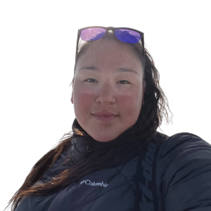 Le français suit. A picture of a person with long dark hair with glasses on her head wearing a black jacket smiling at the camera, framed by a yellow circle.Photo d'une personne aux longs cheveux noirs, portant des lunettes sur la tête et une veste noire, souriant à l'appareil photo, encadrée d'un cercle jaune.