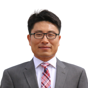 Le français suit. A picture of a person with short black hair wearing glasses and a grey suit smiling at the camera, framed by a green circle.Photo d'une personne aux cheveux courts et noirs, portant des lunettes et un costume gris, souriant à l'appareil photo, encadrée d'un cercle vert.