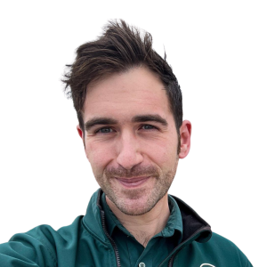 Le français suit. A picture of a person with short brown hair wearing a green shirt smiling at the camera, framed by a yellow circle.Photo d'une personne aux cheveux bruns courts, portant une chemise verte et souriant à l'appareil photo, encadrée d'un cercle jaune.