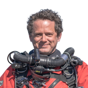 Le français suit. A picture of a person with short grey hair wearing a red diving suit and equipment smiling at the camera, framed by a blue circle.Photo d'une personne aux cheveux gris et courts, portant une combinaison de plongée rouge et un équipement, souriant à l'appareil photo, encadrée d'un cercle bleu.