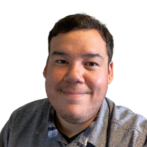 Le français suit. A picture of a person with short black hair wearing a grey shirt smiling at the camera, framed by an orange circle.Photo d'une personne aux cheveux courts et noirs, vêtue d'une chemise grise, souriant à l'appareil photo, encadrée d'un cercle orange.