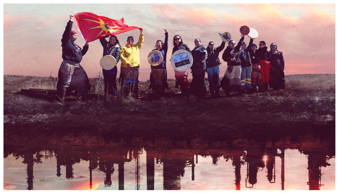 Un groupe de personnes au bord d'une rivière, dont beaucoup portent des jupes à rubans. Certains tiennent des tambours décorés de façon complexe et d'autres brandissent un drapeau des Premières nations. Le reflet dans l'eau est celui d'une usine.