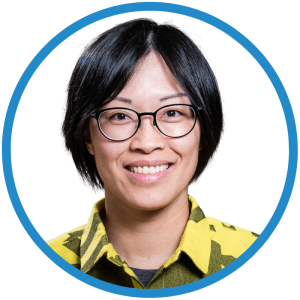 Le français suit A picture of a person with short black hair wearing glasses and a checkered yellow top smiling at the camera, framed by a blue circle. Photo d'une personne aux cheveux courts et noirs, portant des lunettes et un haut jaune à carreaux, souriant à l'appareil photo, encadrée d'un cercle bleu.