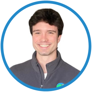 Le français suit. A picture of a person with short black hair wearing a grey sweater smiling at the camera, framed by a blue circle. Photo d'une personne aux cheveux noirs courts, vêtue d'un pull gris, souriant à l'appareil photo, encadrée par un cercle bleu.