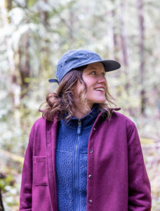 Le français suit. Shauna Doll smiles against a wooded background and looks to the right, a person wearing a pink sweater over a dark blue fleece, with a blue hat and brown hair tied in a bun. Shauna Doll sourit devant un fond boisé et regarde vers la droite, une personne portant un pull rose sur une polaire bleu foncé, avec un chapeau bleu et des cheveux bruns attachés en chignon.