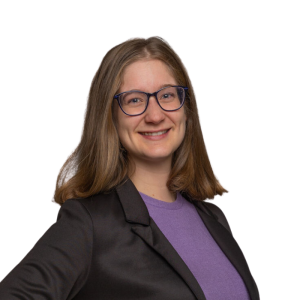 Le français suit<br />
A picture of a person with medium length brown hair wearing glasses and a dark top smiling at the camera, framed by a blue circle.<br />
Photo d'une personne aux cheveux bruns mi-longs, portant des lunettes et un haut sombre, souriant à l'appareil photo, encadrée d'un cercle bleu.<br />
