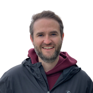Le français suit. A picture of a person with short brown hair wearing a red hoodie and grey jacket smiling at the camera, framed by an orange circle.<br />
Photo d'une personne aux cheveux bruns courts portant un sweat à capuche rouge et une veste grise, souriant à l'appareil photo, encadrée d'un cercle orange.