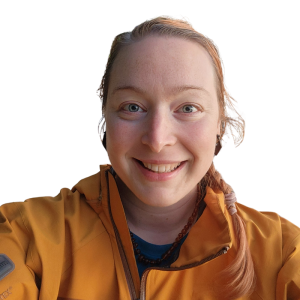 Le français suit.<br />
A picture of a person with long ginger hair wearing an orange jacket smiling at the camera, framed by a green circle.<br />
Photo d'une personne aux longs cheveux roux portant une veste orange et souriant à l'appareil photo, encadrée d'un cercle vert.